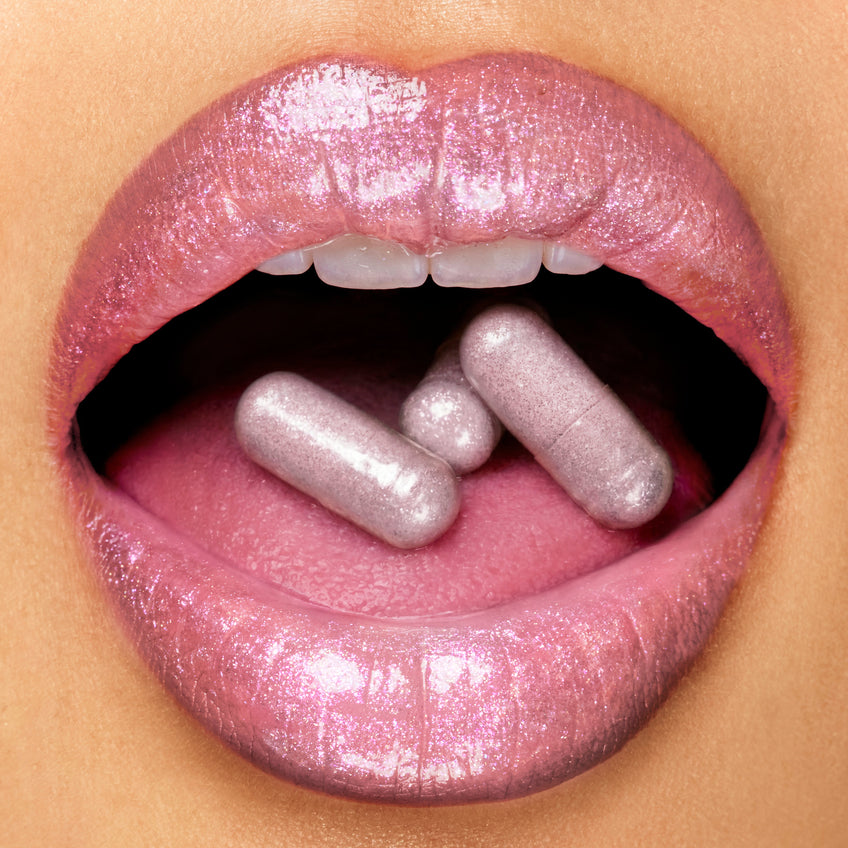 Close-up of a person's mouth with pink lipstick holding silver capsules.